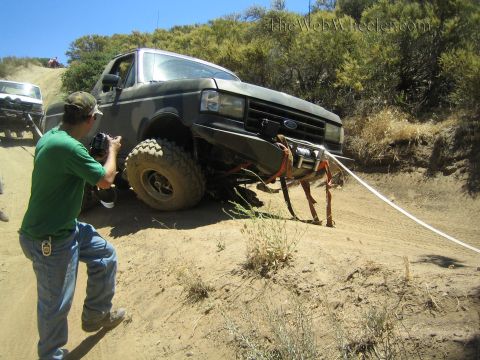 1990 Ford Bronco - man in a green shirt photographs a dark off-road pickup with a damaged front suspension perched on a sandy hillside trail.