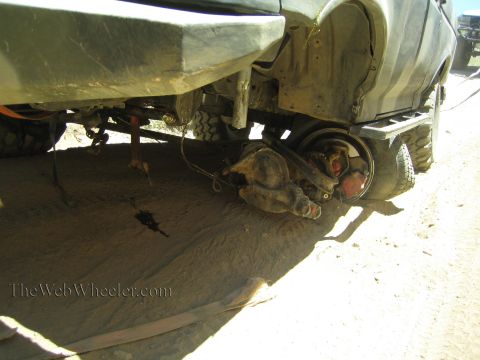 1990 Ford Bronco - Underbody view of an off-road truck showing the front axle, brake hub and drivetrain components over sandy ground.