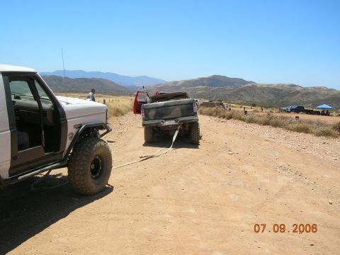 1985 Ford Bronco - Two off-road vehicles are on a dirt road, one towing the other, with a mountainous landscape under a clear blue sky.