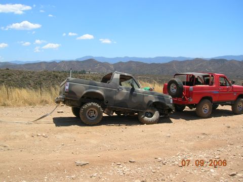 1985 Ford Bronco - Two Ford Broncos, one camouflaged and the other red, are seen in a recovery scene on a dirt road with mountains in the background.