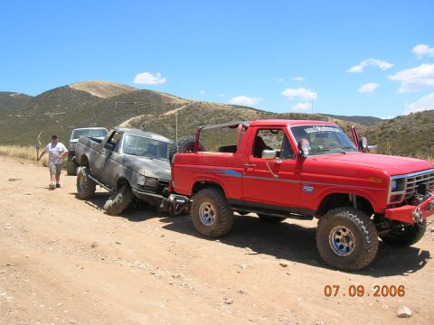 1985 Ford Bronco - A red Ford Bronco is assisting a stuck gray Ford pickup truck on a dirt road with hills in the background.