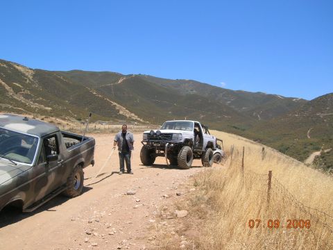 1985 Ford Bronco - Two off-road trucks are on a dirt road in a dry, hilly area. A man is holding a shovel near the white truck.