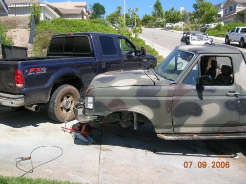 1985 Ford Bronco - A camouflaged pickup truck is being worked on, with a dark blue Ford F-series truck parked beside it on a sunny day.