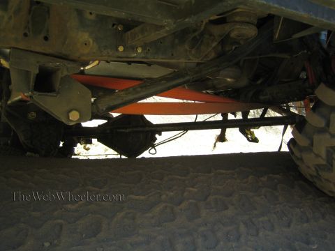1990 Ford Bronco - Underbody of a lifted off-road truck showing the frame rails, axle and an orange-red recovery strap running across.