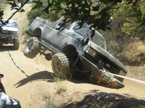 1990 Ford Bronco - camo Ford pickup with a front winch tilts steeply on an eroded dirt trail, another off-road truck behind it.