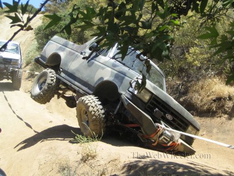 1990 Ford Bronco - camo Ford pickup with a front winch leans at a sharp angle on a dirt off-road trail beneath overhanging branches.