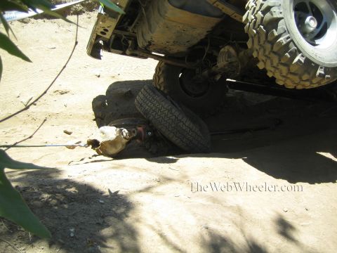1990 Ford Bronco - Underbody of an off-road truck with a wheel and damaged front axle components lying on the sandy ground beneath it.