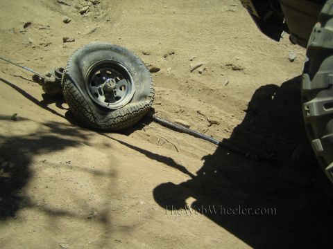 1990 Ford Bronco - wheel and a broken axle shaft lie on the dirt next to a large off-road tire, a person's shadow cast nearby.