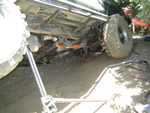 1990 Ford Bronco - Underbody of a lifted off-road truck raised on a high-lift jack with tools on the ground during a trail-side repair.