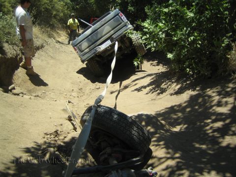 1990 Ford Bronco - light-colored pickup tilts on a dirt off-road trail while a detached wheel and recovery strap sit on the ground ahead.