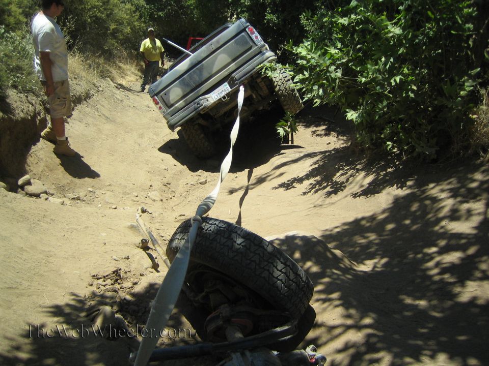 Light pickup tilted on a dirt trail with a detached wheel and strap in front
