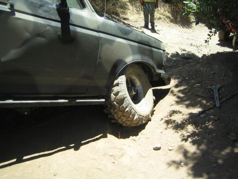 1990 Ford Bronco - front off-road tire and wheel of a dark truck sit lodged in a sandy rut on a shaded trail, a wrench on the ground.