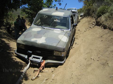 1990 Ford Bronco - camo-painted Ford pickup with a front winch sits on a sandy off-road trail as people gather behind it.