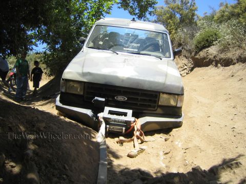 1990 Ford Bronco - white Ford pickup with a front winch sits on a rutted dirt trail as two people walk along the shaded path behind.