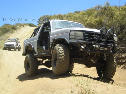 1990 Ford Bronco - lifted silver Ford Bronco on oversized mud tires crawls up a dusty off-road hill, with another rig behind it.