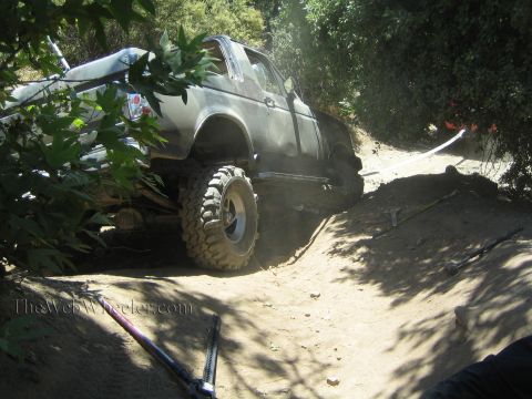 1990 Ford Bronco - lifted open-cab truck on mud tires articulates over a steep dirt obstacle in dappled shade, raising dust.