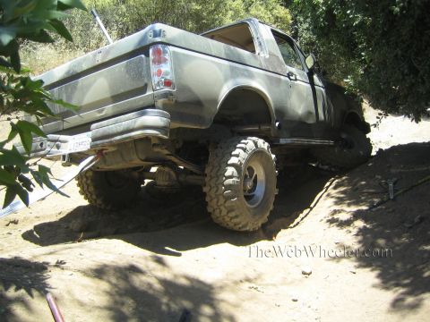 1990 Ford Bronco - rear of a lifted, stripped pickup on large mud tires sits on a dirt trail surrounded by green brush.