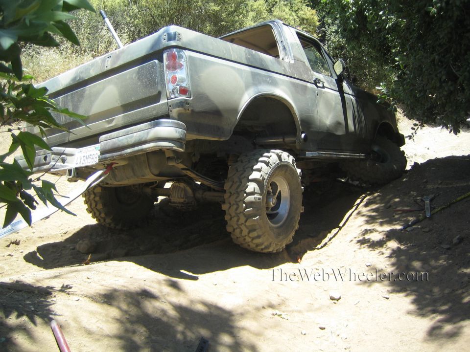 Rear view of a lifted bare-metal pickup on mud tires parked on a shaded dirt trail