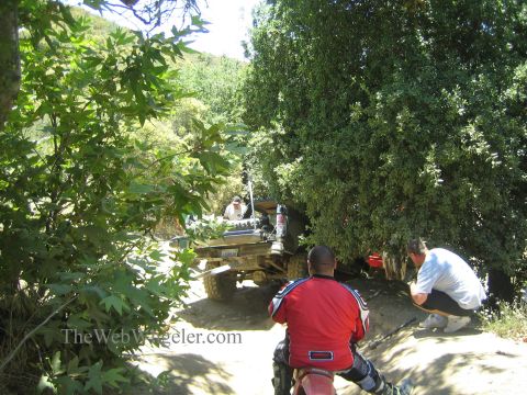 1990 Ford Bronco - Two onlookers, one kneeling in a red jacket, watch a small off-road vehicle parked beneath shady trees on a trail.