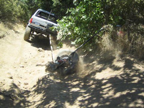 1990 Ford Bronco - lifted SUV climbs a dusty hill while a broken axle assembly and wheel drag behind it on a yellow strap.