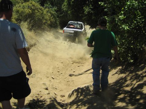 1990 Ford Bronco - Two bystanders watch from the trailside as a pickup kicks up dust speeding up a rutted dirt hill.