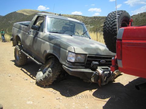 1990 Ford Bronco - camo-painted lifted Ford pickup with a front winch bumper sits on a dirt lot beside a red truck in dry hills.