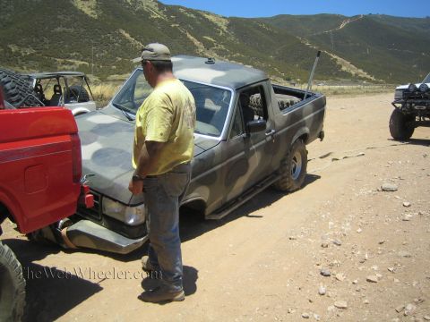 1990 Ford Bronco - man in a yellow shirt and cap stands next to a muddy camo-painted Ford pickup parked on a dirt lot in dry hills.