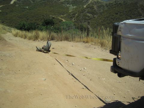 1990 Ford Bronco - broken-off axle with a wheel lies on a dirt trail, connected by a yellow strap to a vehicle at right.