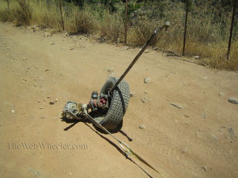 1990 Ford Bronco - complete detached axle assembly with tire and driveshaft rests on a dirt road beside a wire fence.