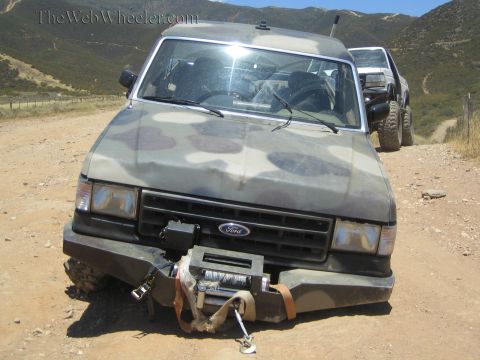 1990 Ford Bronco - Close front view of a camo-painted Ford truck with a collapsed front end and winch dangling from the bumper.