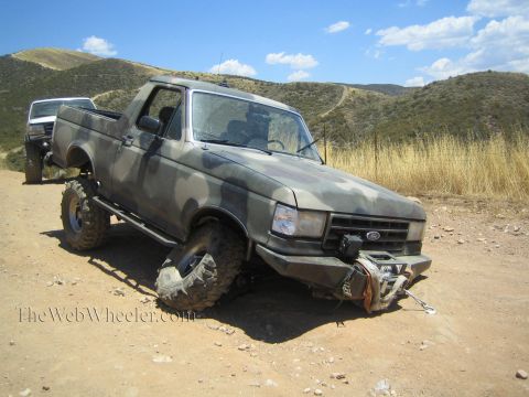 1990 Ford Bronco - camo-painted lifted Ford pickup sits in dry hills with its front wheel splayed out from a broken axle.