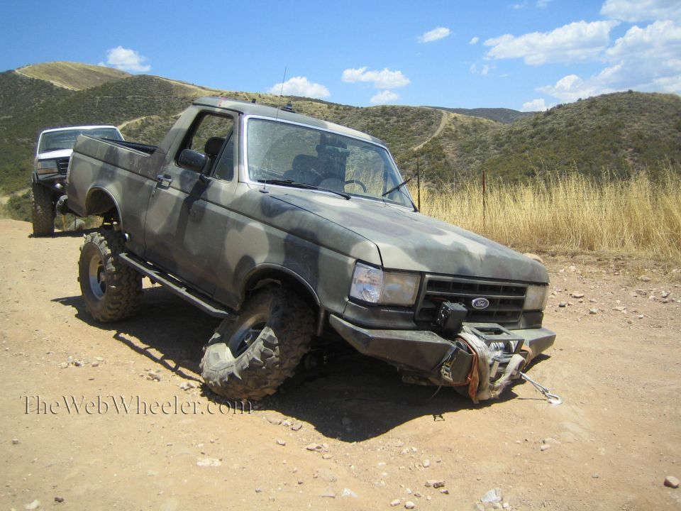 Camo lifted Ford pickup with broken front wheel sitting on a dirt lot in dry hills