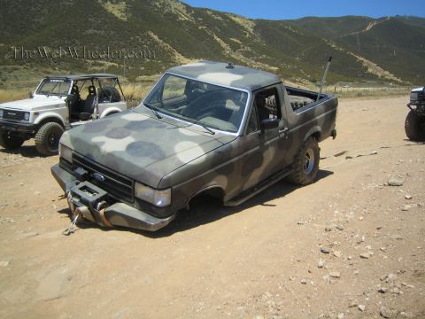 1990 Ford Bronco - camo-painted Ford pickup sits low on a collapsed front end on a dirt lot, with other off-road rigs nearby.