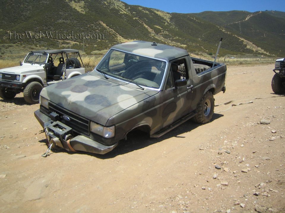 Camo Ford pickup with collapsed front suspension parked on a dirt lot near other off-road rigs