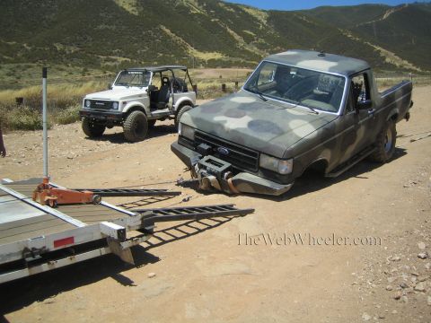 1990 Ford Bronco - camo Ford pickup with a broken front end sits beside an aluminum car trailer with ramps deployed on a dirt lot.