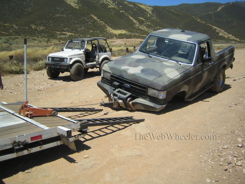 Camo Ford pickup with broken front end next to an aluminum trailer with ramps on a dirt lot