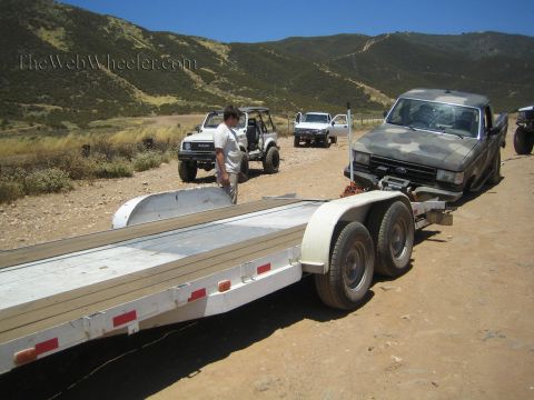 1990 Ford Bronco - empty aluminum tandem-axle trailer is positioned near a camo Ford truck while people gather on a dirt lot in dry hills.