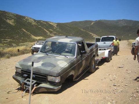 1990 Ford Bronco - camo-painted Ford truck is staged for loading onto an aluminum trailer on a dirt lot, with people standing nearby.