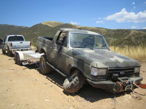 1990 Ford Bronco - camo Ford truck with a splayed broken front wheel sits beside a trailer hitched to a white dually pickup.