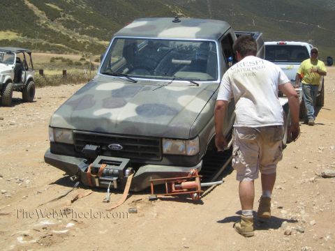 1990 Ford Bronco - man in a white shirt and shorts walks toward a camo-painted Ford SUV sitting on a collapsed front end on a dirt lot.