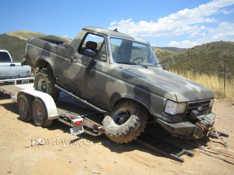 1990 Ford Bronco - camo-painted Ford truck with a splayed front wheel sits loaded on an aluminum tandem-axle trailer in dry hills.