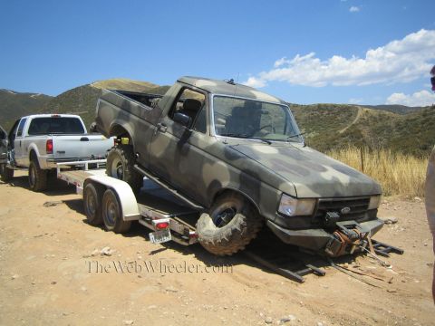 1990 Ford Bronco - camo-painted Ford truck with a broken front wheel rides on an aluminum trailer behind a white dually in dry hills.