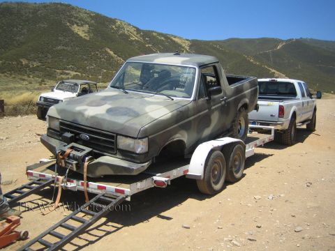 1990 Ford Bronco - camo-painted Ford truck sits loaded on a tandem-axle trailer hitched to a white dually pickup on a dirt lot.