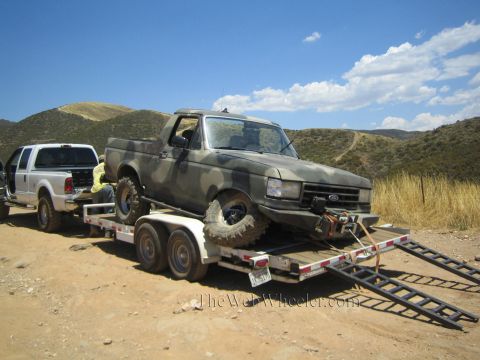 1990 Ford Bronco - camo Ford truck with a splayed front wheel sits on a trailer with ramps down, hitched to a white dually in the hills.