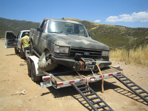 1990 Ford Bronco - Front view of a muddy Ford pickup loaded on a trailer with ramps down, a person in yellow standing near the open truck door.