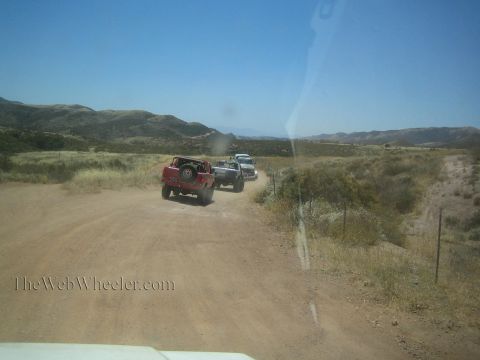 1990 Ford Bronco - red truck and a white truck travel along a dusty dirt road winding through arid scrubland and rolling hills.