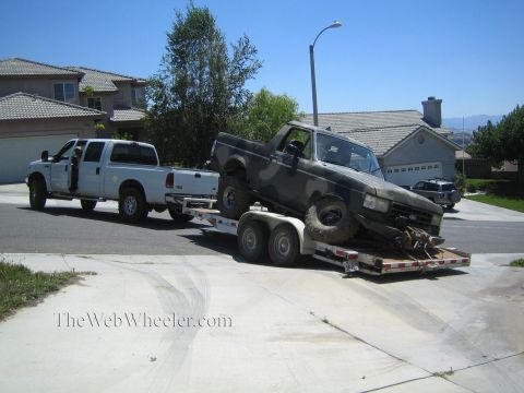 1990 Ford Bronco - camo-painted Ford pickup with large off-road tires sits on a tandem-axle flatbed trailer behind a white crew-cab truck in a suburban drivew…
