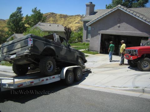 1990 Ford Bronco - camouflage-painted Ford SUV body sits on a trailer in a driveway while two men stand near an open garage with a red lifted truck nearby.