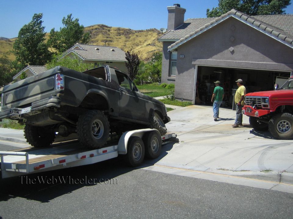 Camo Ford SUV on a flatbed trailer in a driveway with two men by a garage