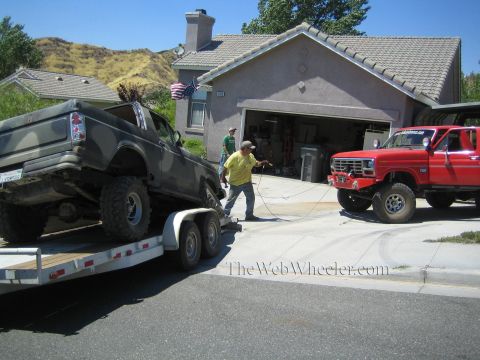 1990 Ford Bronco - man in a yellow shirt works in a driveway between a camo-painted truck on a trailer and a lifted red Ford pickup outside a tile-roofed hous…